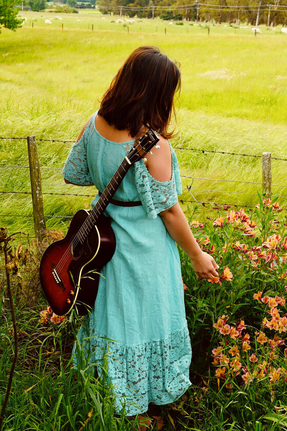 Katrina Lalo Louise walking a fence line with her guitar, reaching for wildflowers
