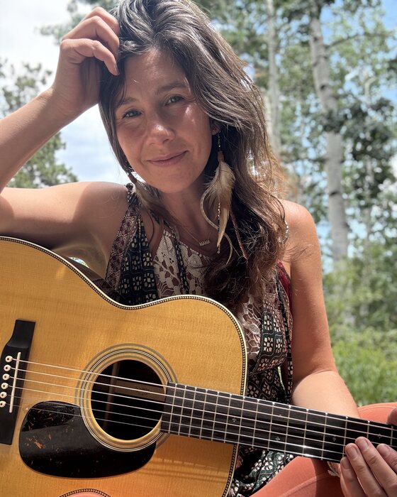 Katrina Lalo Louise outdoors with her guitar, aspens behind her