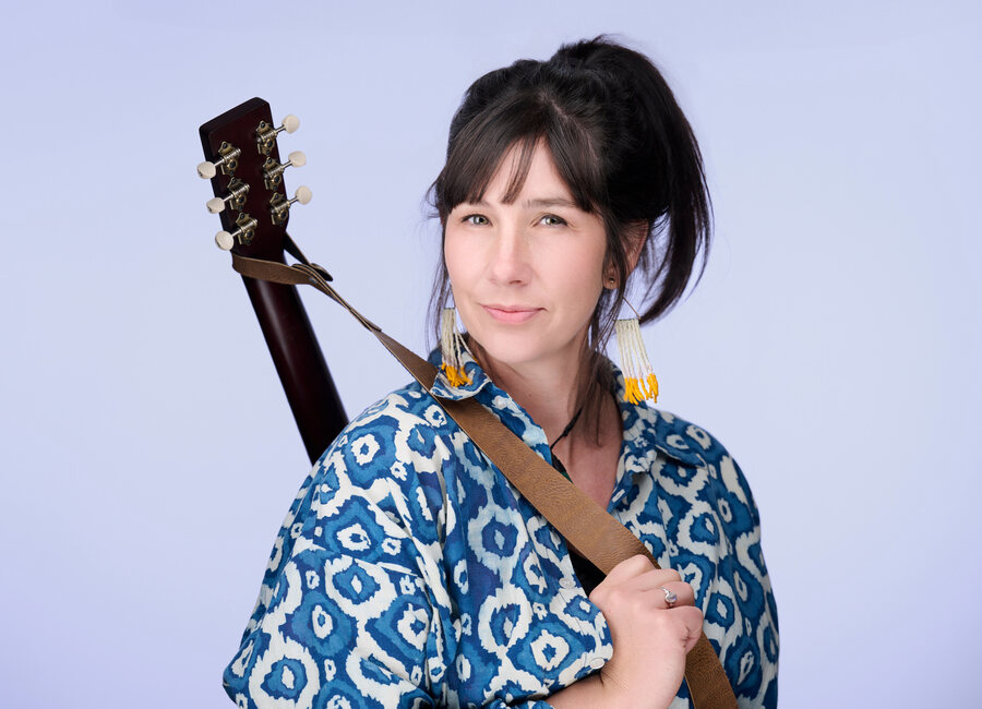 Katrina Lalo Louise standing with her guitar on its strap, against a plain backdrop