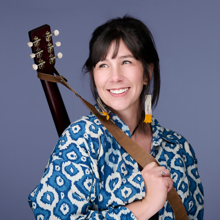 Katrina Lalo Louise with her guitar over one shoulder, against a plain backdrop